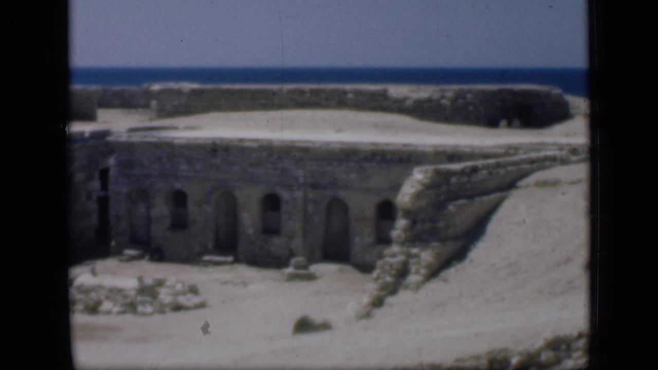 1946 Egypt home-movie frame with thick masonry walls, repeated arched doorways, sandy open ground, and the sea visible beyond.