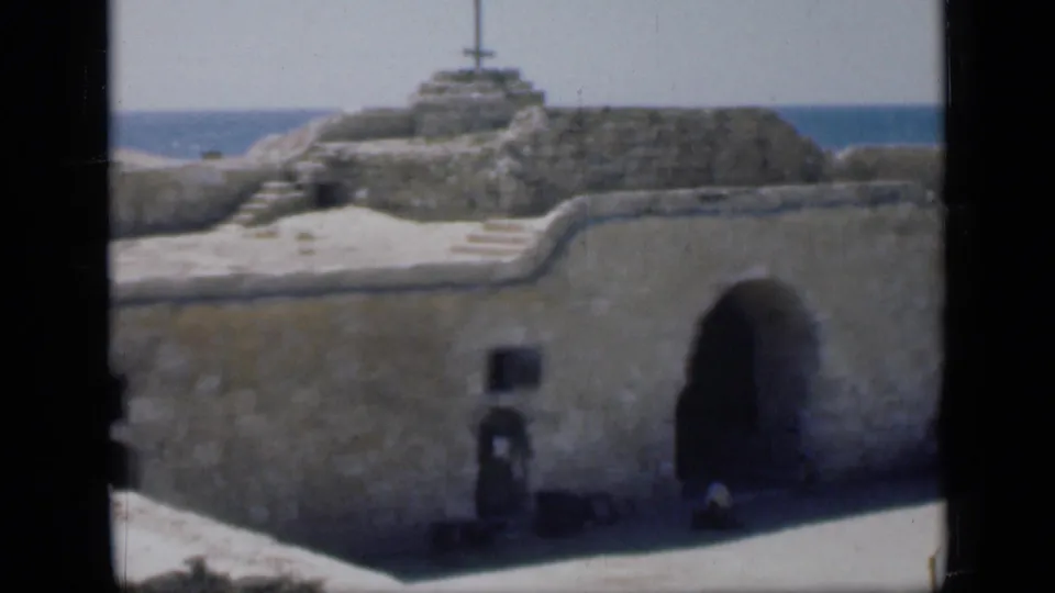 1946 Egypt home-movie frame showing a large arched gateway, thick stone wall, steps, and a cross-topped mound near the sea.