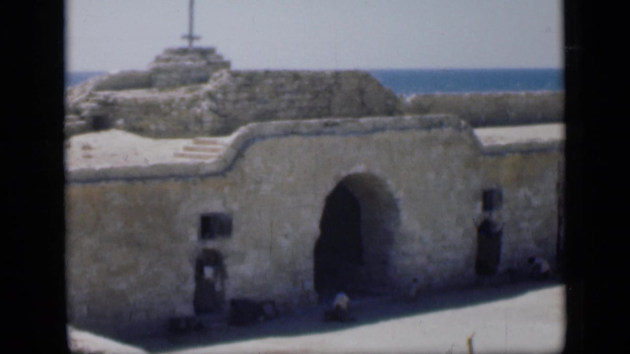 Stone ruin facade with arched opening, small windows, raised section, and blue water visible above the wall.