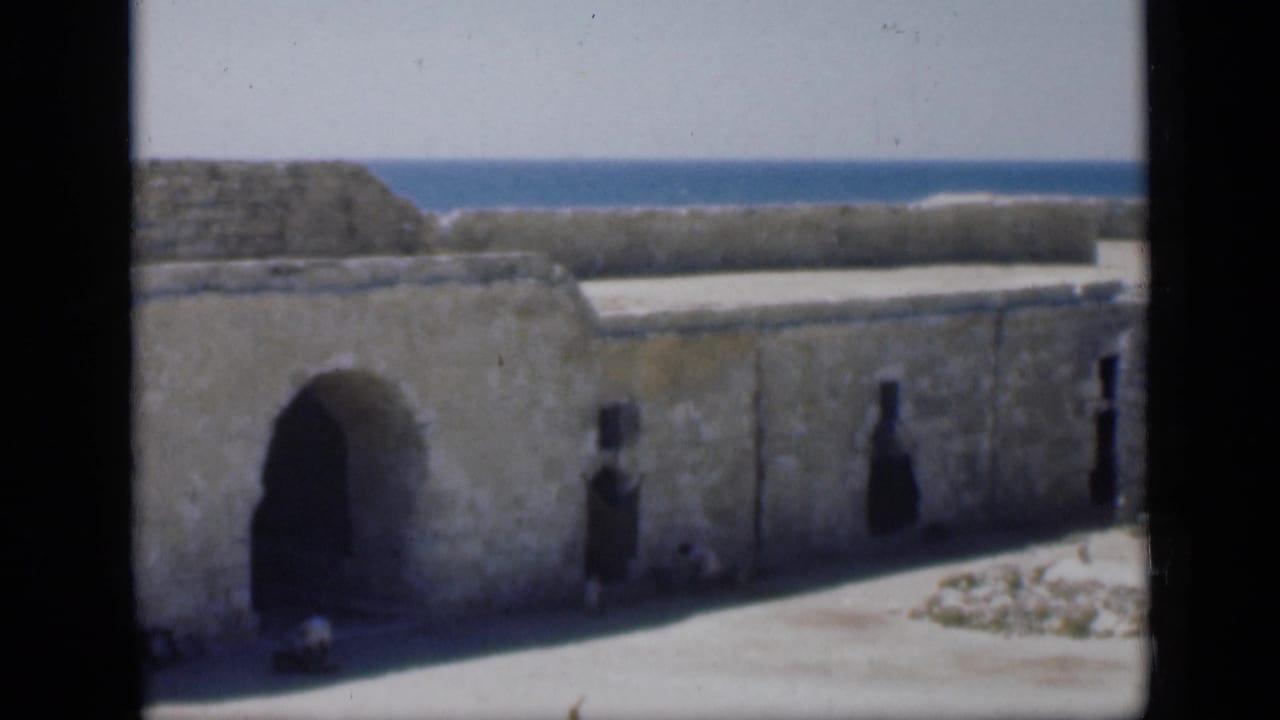 1946 Egypt 8mm frame showing a weathered stone fort wall, arched entrance, sunlit ground, and blue sea beyond.