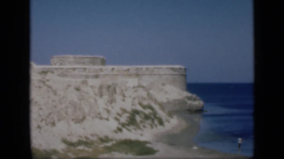1946 Egypt 8mm frame showing a broad stone fort wall on a coastal rise, with rock, vegetation, and deep blue water nearby.