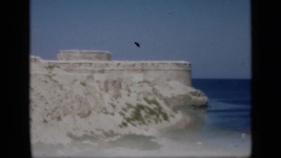 1946 Egypt frame showing the coastal fortress, rocky slope, blue sea, and a small distant visitor near the shoreline.