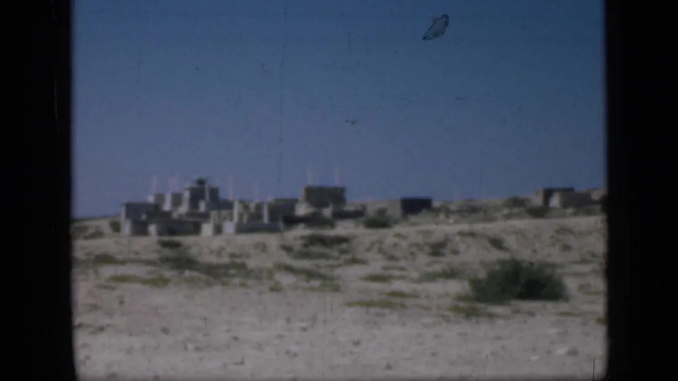 1946 Cairo-area frame with grainy 8mm texture, sandy foreground, sparse plants, and distant low stone buildings.