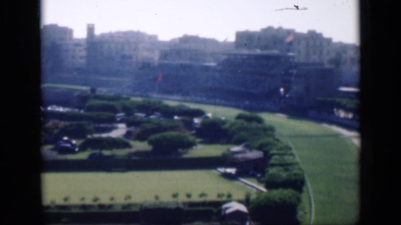 1946 Cairo home-movie frame showing manicured garden grounds with lawns, hedges, parked cars, and pale urban buildings in the distance.