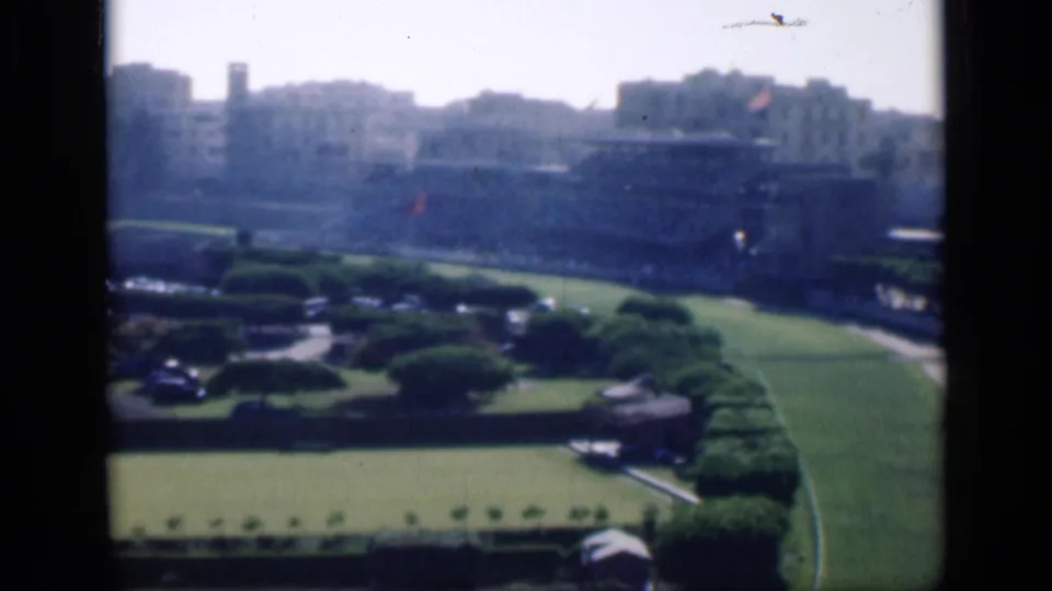 1946 Cairo home-movie frame showing manicured garden grounds with lawns, hedges, parked cars, and pale urban buildings in the distance.