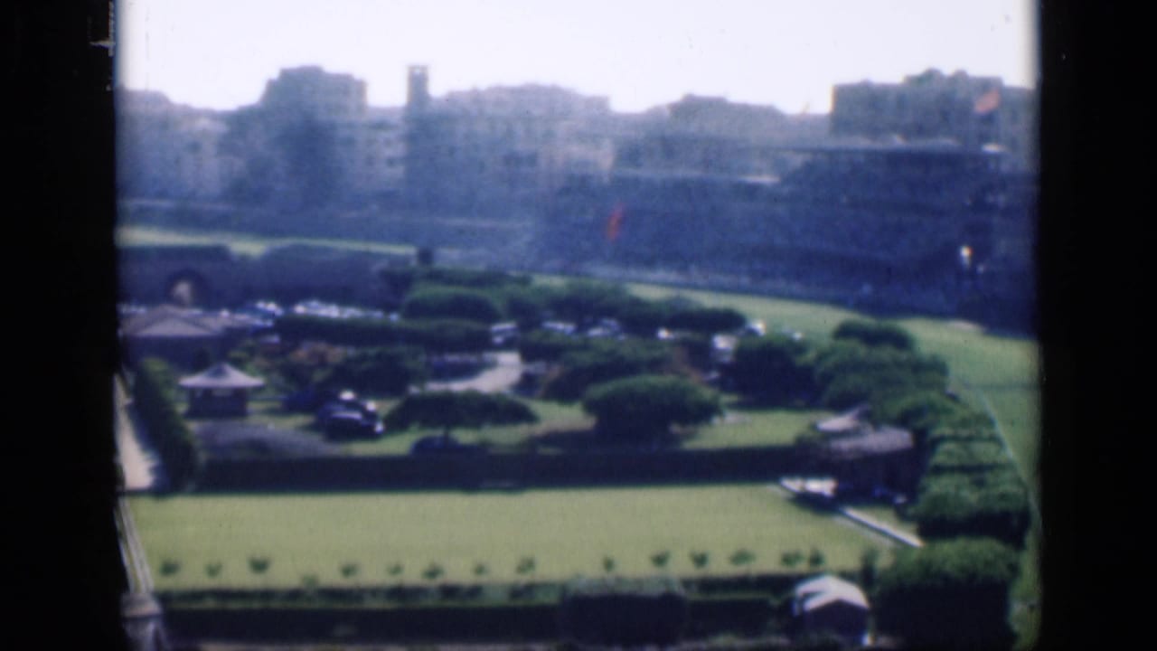 Formal garden grounds with hedges, lawns, a small pavilion-like structure, parked cars, and city buildings.