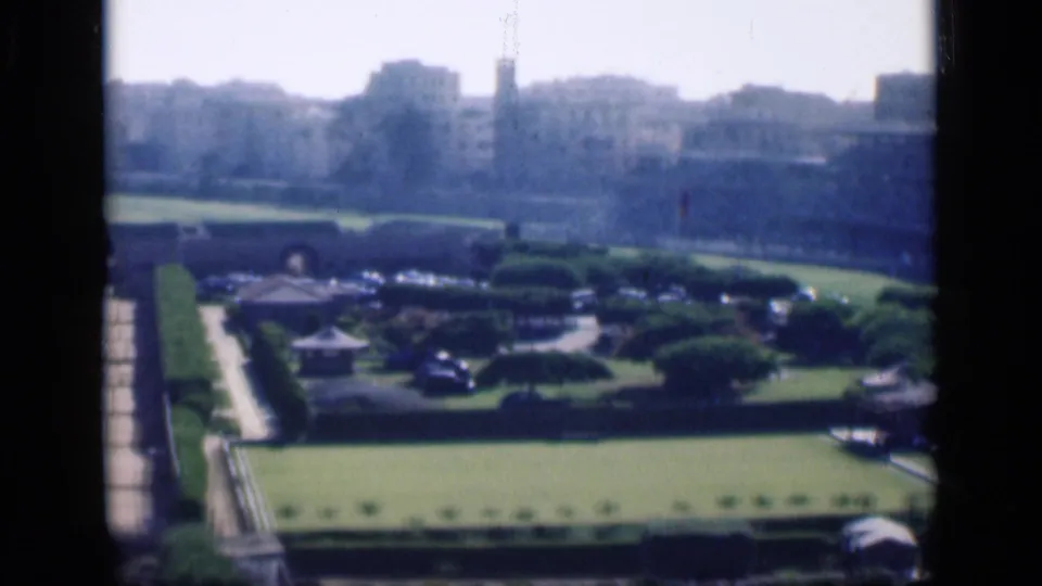 1946 Cairo home-movie view over formal lawns, a broad paved approach, clipped hedges, cars, and distant urban buildings.