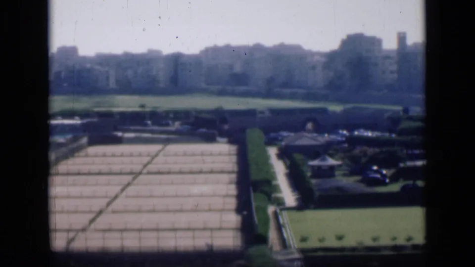 Elevated 1946 Cairo view with gridded farm plots, manicured hedges, small structures, parked vehicles, and a soft city skyline.