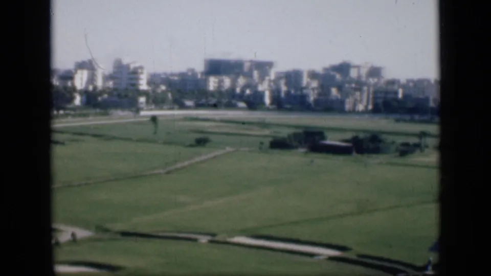 1946 Cairo home-movie frame with a broad green field, pale walkways, small distant figures, and urban buildings beyond.