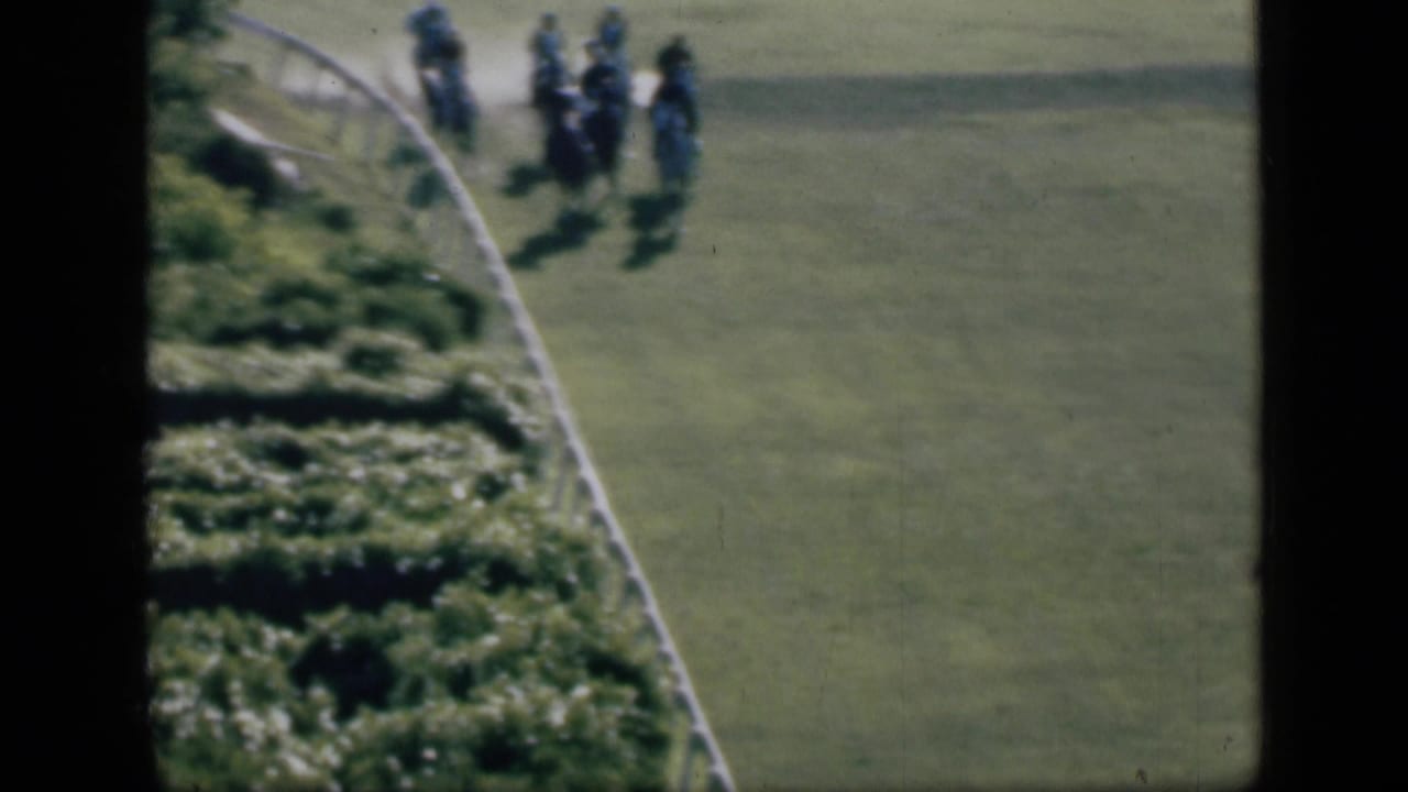 Small group of jockeys riding horses around a curved green track beside a white rail.