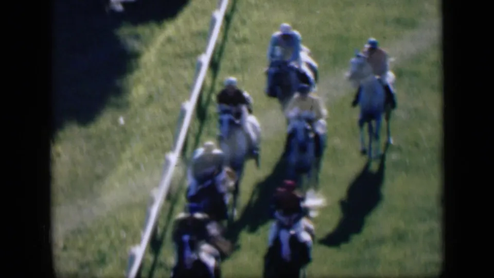 Close high-angle 8mm frame of jockeys in muted silks riding tightly grouped horses beside the track rail in Cairo.