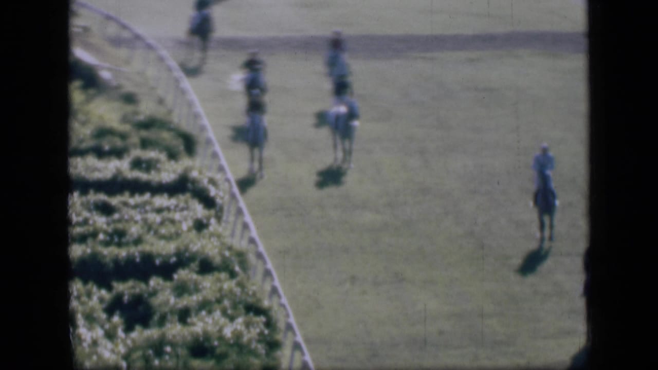 Distant jockeys on horseback walk along a grassy racetrack beside a white rail.