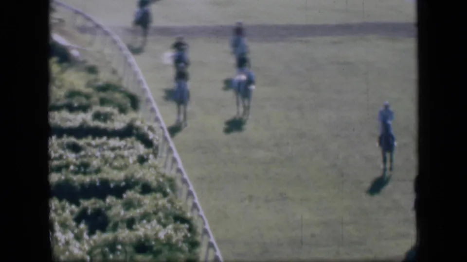 High-angle 1946 Cairo home-movie view of several jockeys walking horses on a grass racetrack near a curved white fence.