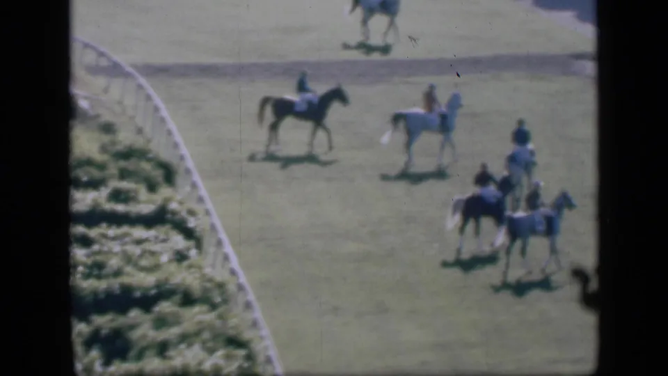 1946 Cairo home-movie frame showing riders on dark and light horses crossing a grassy racetrack near a white fence.