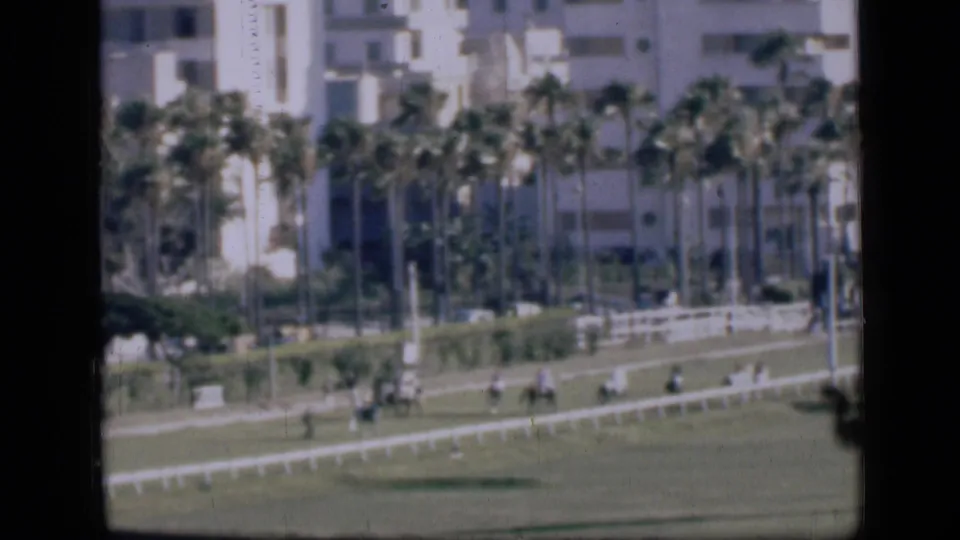 Wide 1946 Cairo frame showing distant riders on a racetrack with white fencing, palm trees, and urban buildings behind.