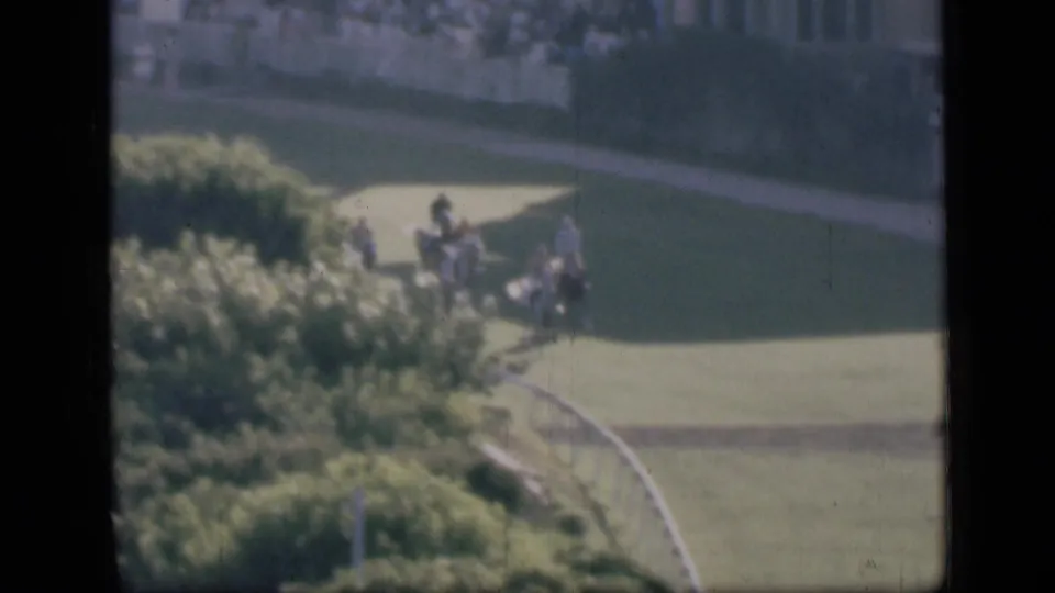 Home-movie frame from the Cairo racetrack showing a small group of people and horses on grass beside shrubs and a curved white rail.