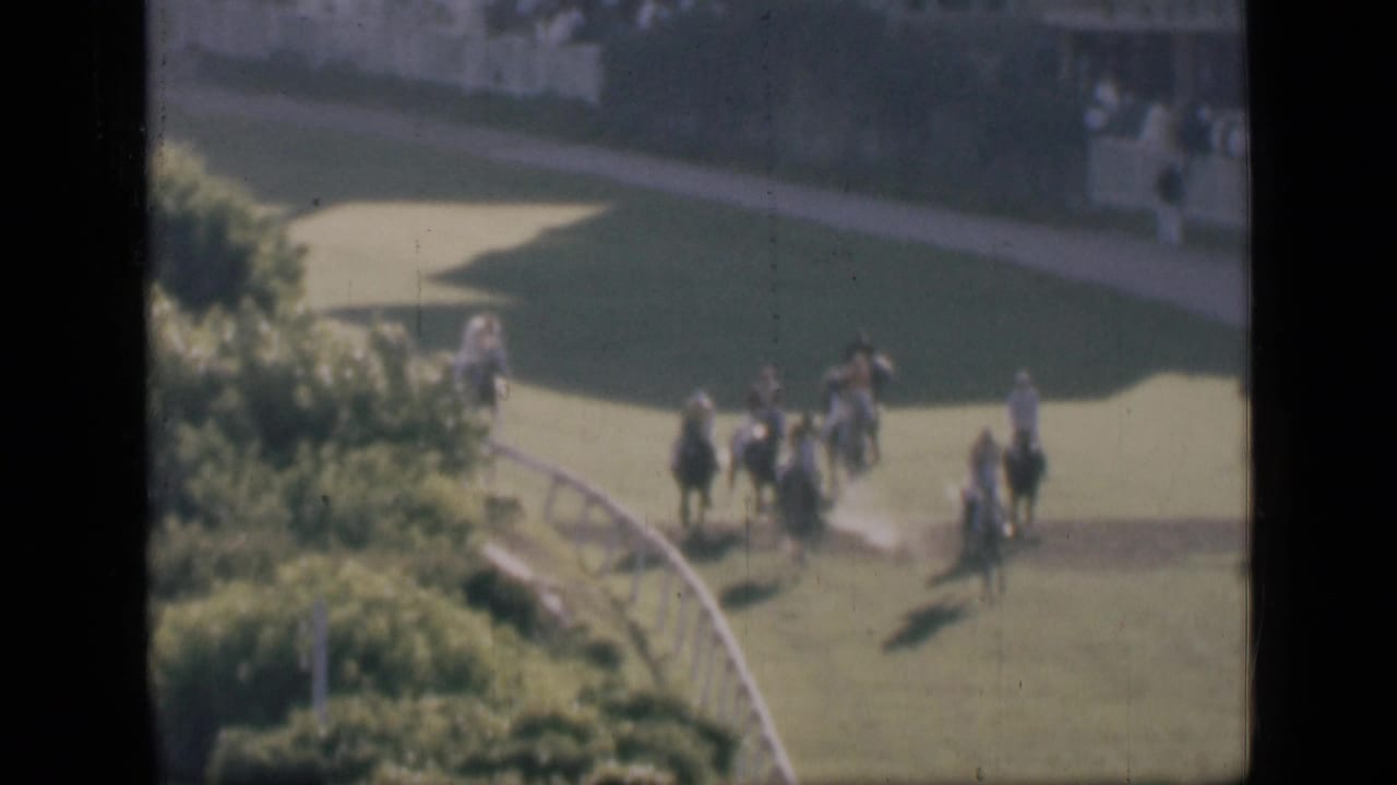 1946 Cairo home-movie frame showing horse riders rounding a green racecourse bend, with spectators in the background.