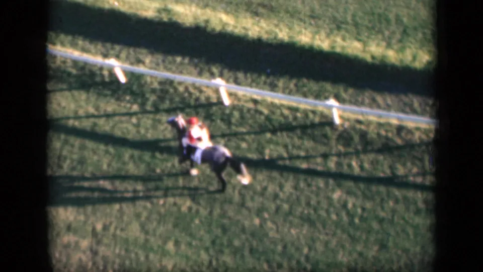 1946 Cairo home-movie frame of a jockey in red and white silks riding a dark horse across green turf beside white rail fencing.