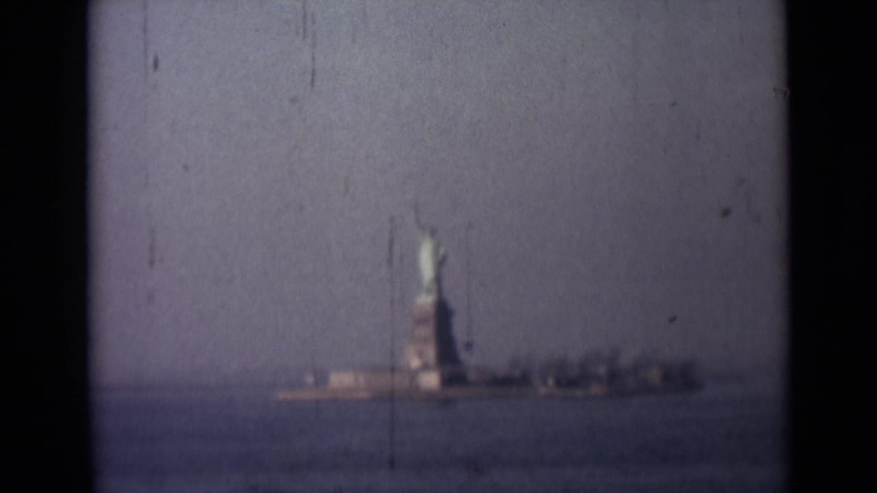 Soft 1940s home-movie frame of the Statue of Liberty across water, with low buildings and heavy film wear visible.