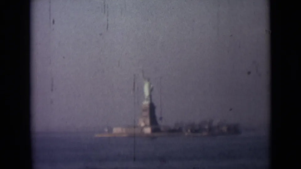 Soft 1940s home-movie frame of the Statue of Liberty across water, with low buildings and heavy film wear visible.