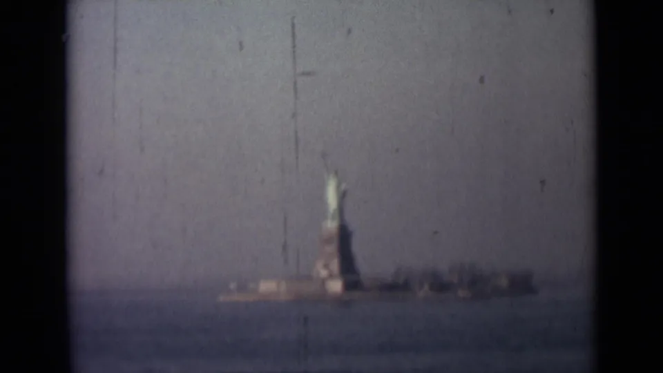 1940s 8mm still with the Statue of Liberty centered low in frame, seen across water through haze and visible scratches.