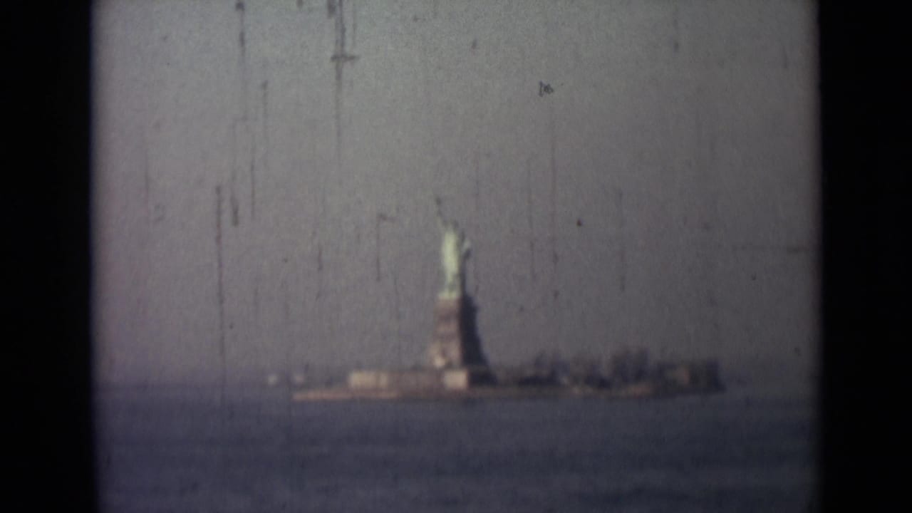 Distant 1940s 8mm harbor view of the Statue of Liberty, with soft focus, muted color, and film scratches visible.