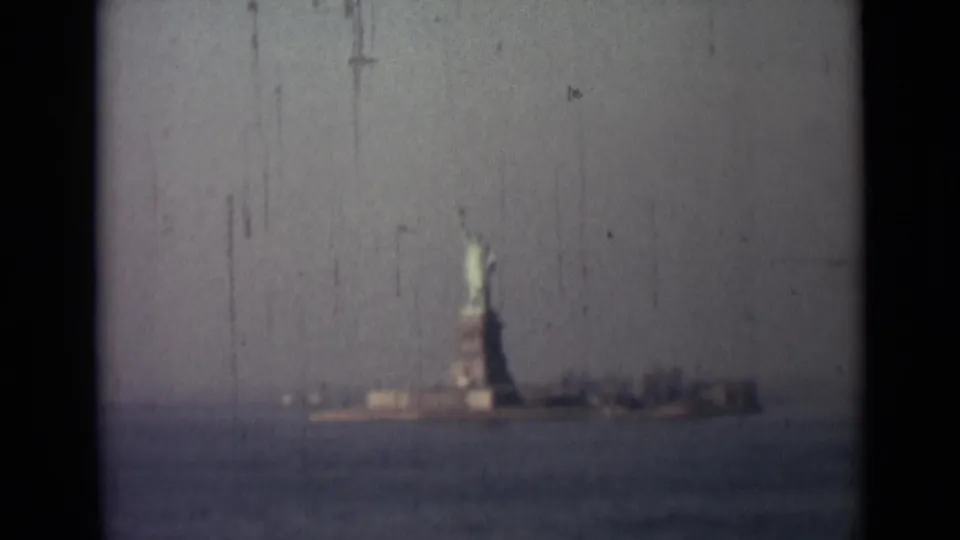 Distant 1940s 8mm harbor view of the Statue of Liberty, with soft focus, muted color, and film scratches visible.