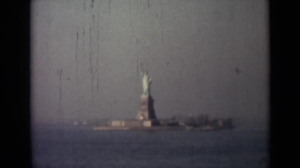 Distant 1940s 8mm view of the Statue of Liberty on Liberty Island, with water foreground and visible gate border.