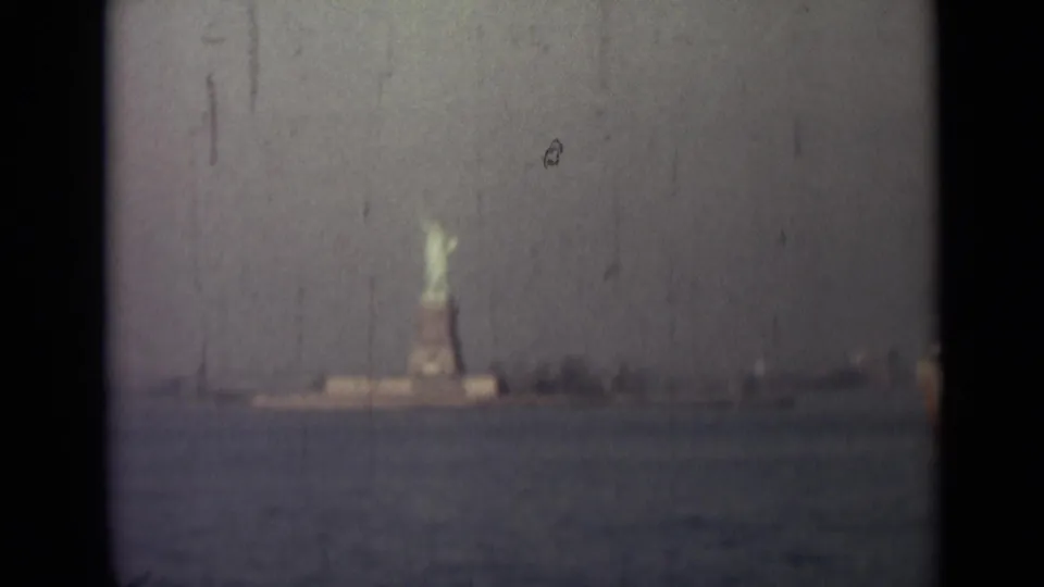 Distant 8mm view of the Statue of Liberty and pedestal across harbor water, with soft focus, dust, scratches, and muted color.