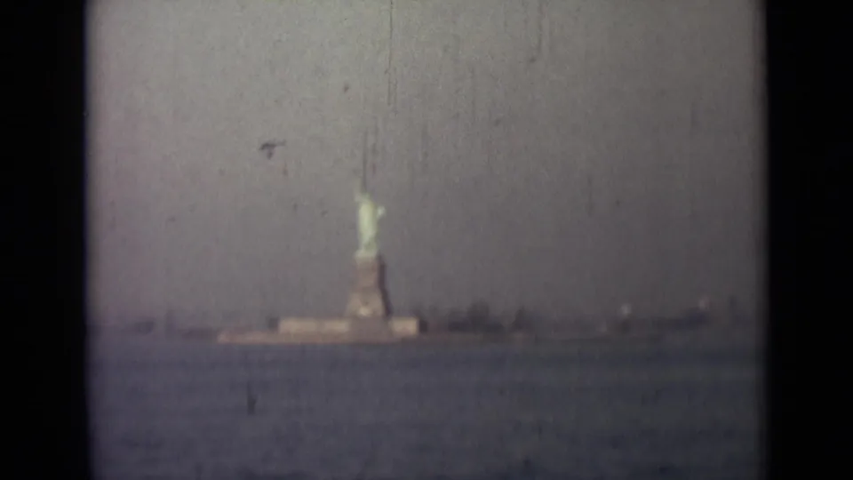 Soft 1940s 8mm frame showing the Statue of Liberty from a distance, with open water, shoreline structures, and visible film aging.