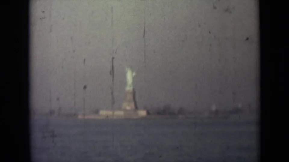 Muted 8mm home-movie view of the Statue of Liberty across water, framed by dark gate edges and marked by dust and vertical scratches.