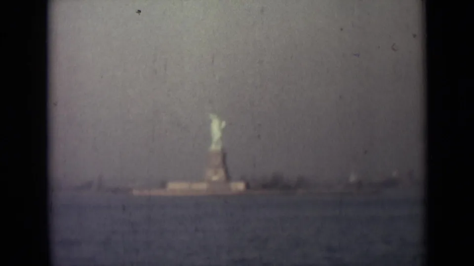 Distant Statue of Liberty seen across water in archival 8mm footage, with hazy sky, faint shoreline, and visible film wear.