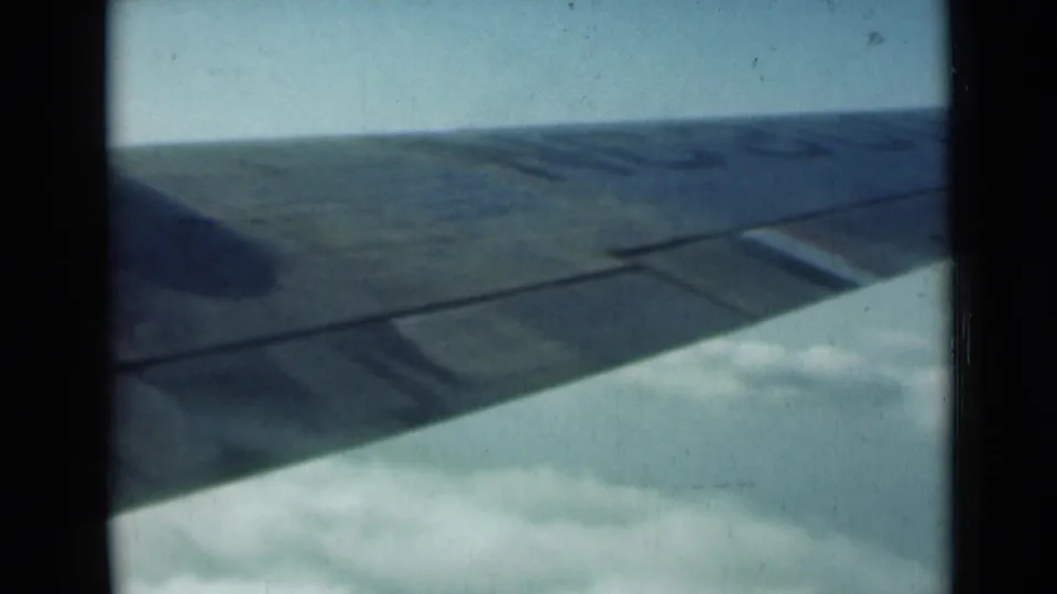 Passenger-window view of an airplane wing above clouds in faded 8mm color footage from a 1940s travel sequence.