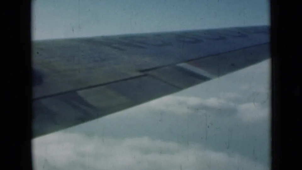 A broad airplane wing fills the passenger-window view over clouds, with muted color and visible 8mm film wear.