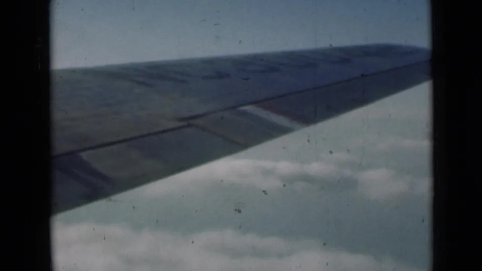 Faded 8mm passenger-window footage shows an airplane wing angled over clouds, with scratches and dust visible in the frame.