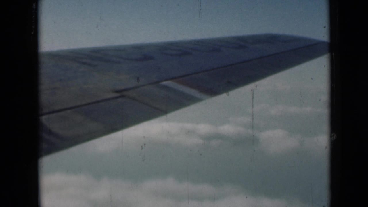 1946 London-context home-movie frame showing an airplane wing extending over pale clouds and blue sky.
