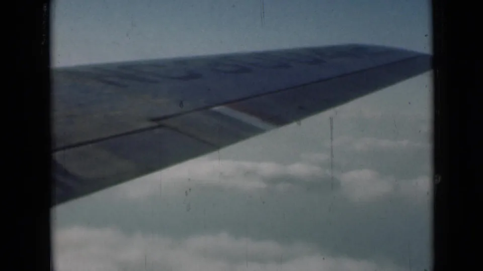 1946 London-context home-movie frame showing an airplane wing extending over pale clouds and blue sky.