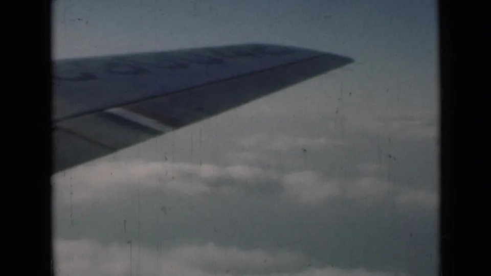 8mm frame showing the airplane wing lower in the view, with clouds below and archival scratches visible.
