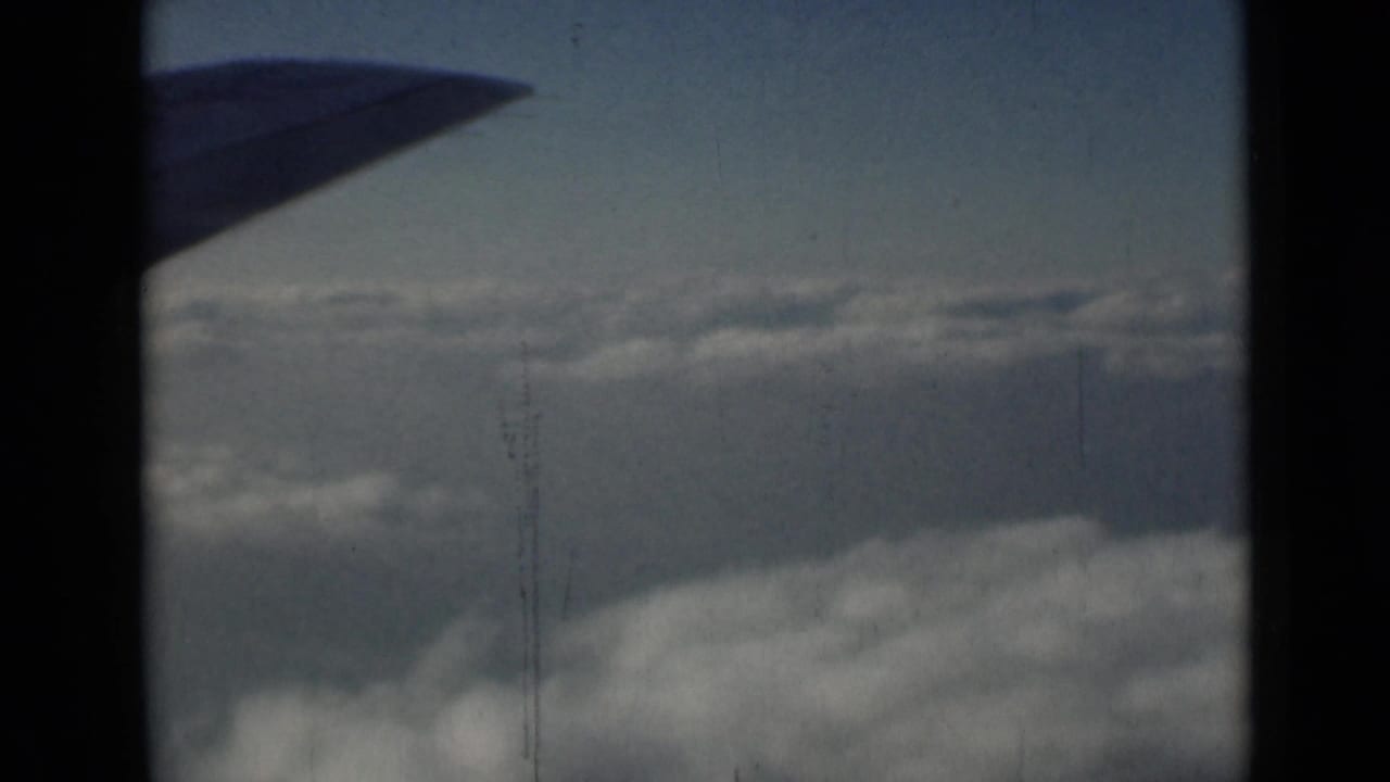 Passenger-window view of a silhouetted aircraft wing over clouds, with 8mm grain and faint vertical wear marks.