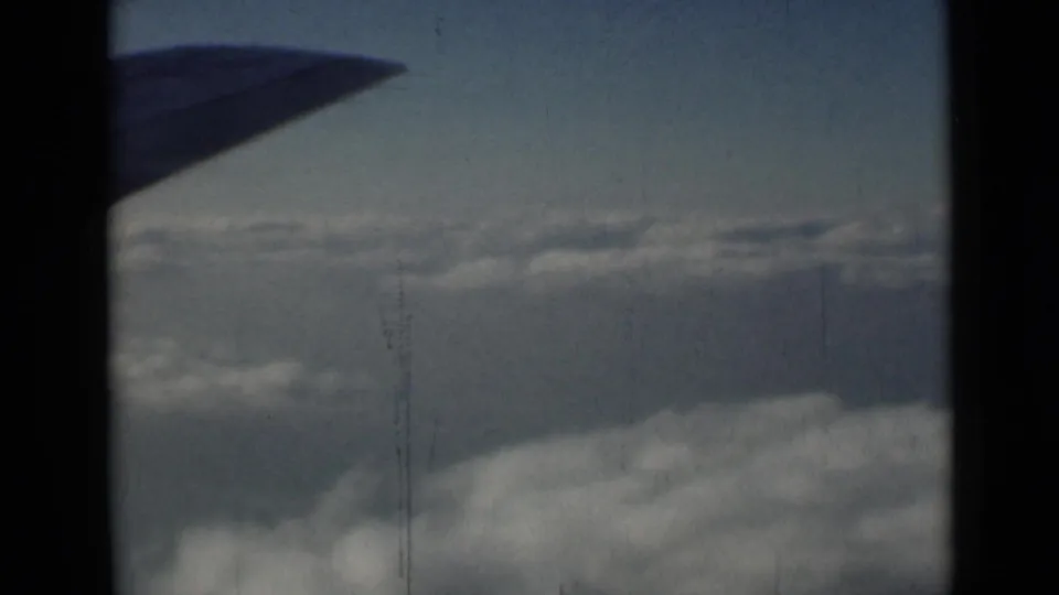 Passenger-window view of a silhouetted aircraft wing over clouds, with 8mm grain and faint vertical wear marks.