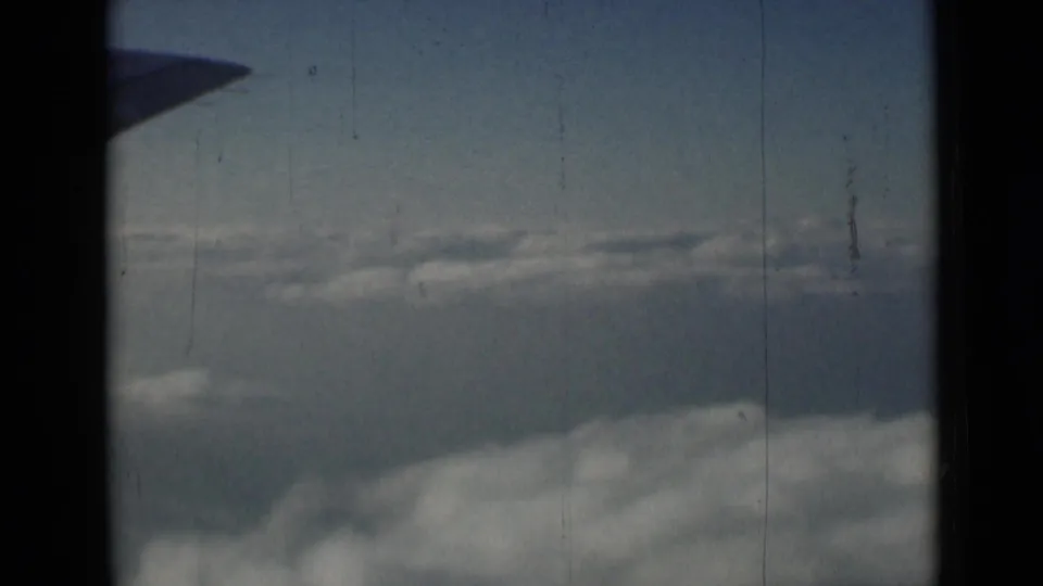 A partial wing edge enters the upper frame as layered clouds pass below in archival 8mm travel footage.