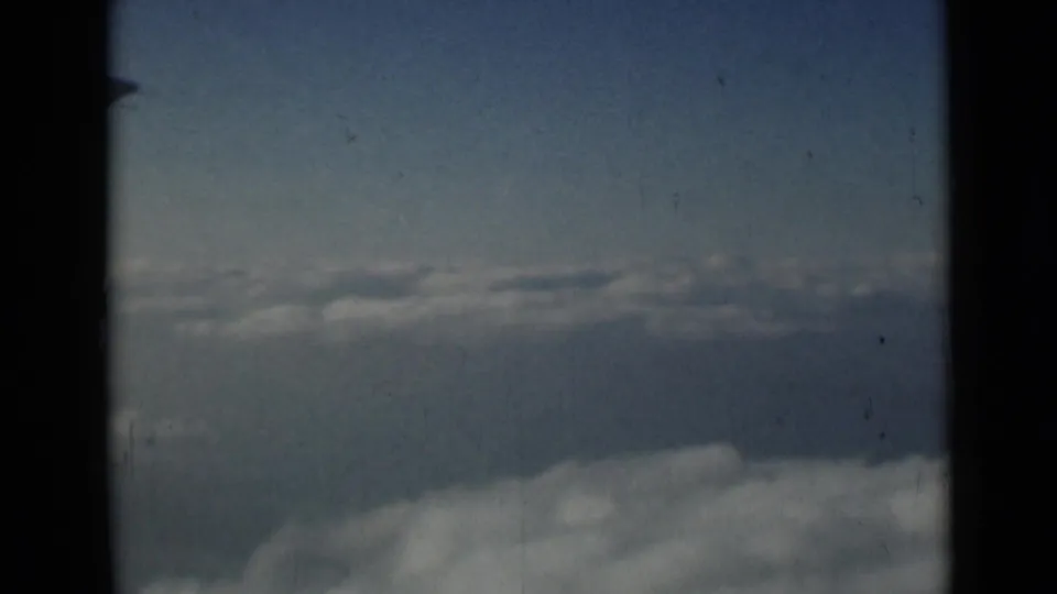 Fixed airplane-window view of clouds and pale sky, with a small wing edge and visible 8mm film wear.