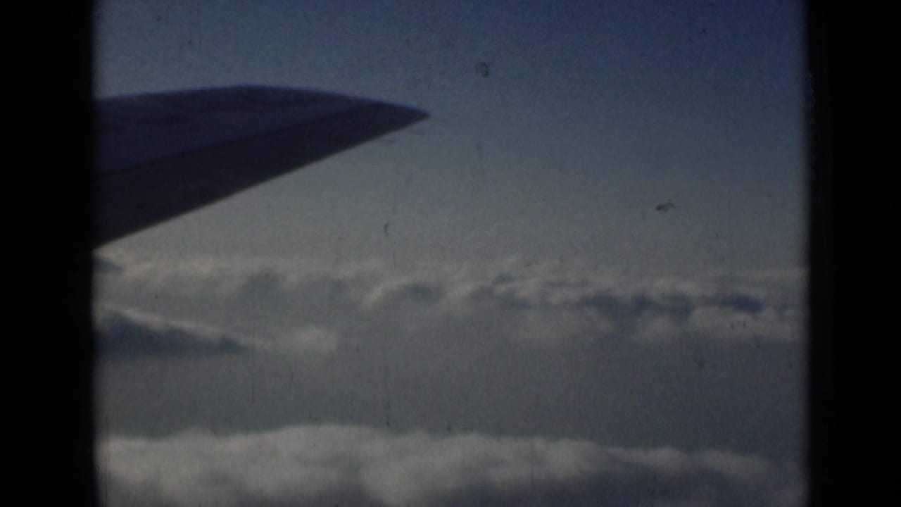 1946 8mm frame showing a dark airplane wing over clouds, with blue sky and visible film scratches.