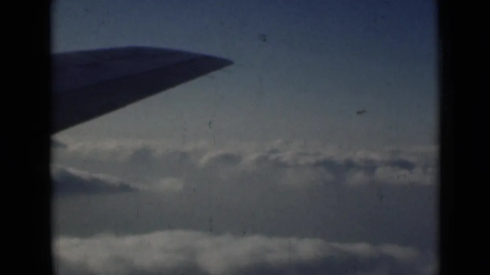 1946 8mm frame showing a dark airplane wing over clouds, with blue sky and visible film scratches.