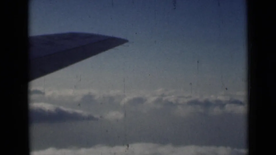 1946 8mm frame of an airplane wing over clouds, with muted color, scratches, and dark film borders.