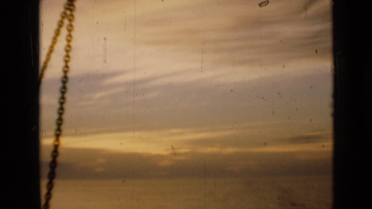 1946 London home-movie frame of a warm sunset sky viewed through a window, with a dark vertical edge and hanging chain in the foreground.