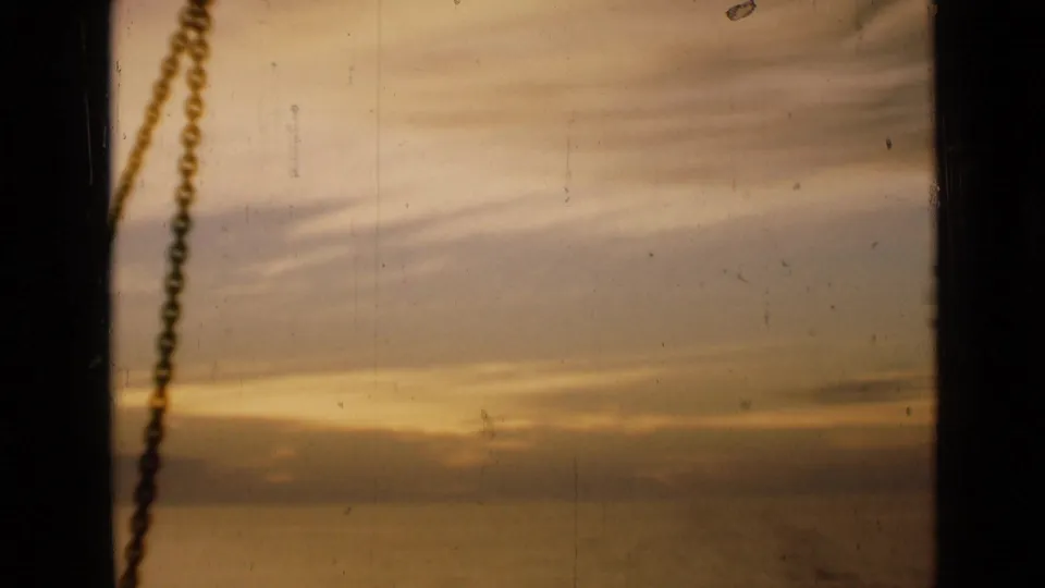 1946 London home-movie frame of a warm sunset sky viewed through a window, with a dark vertical edge and hanging chain in the foreground.