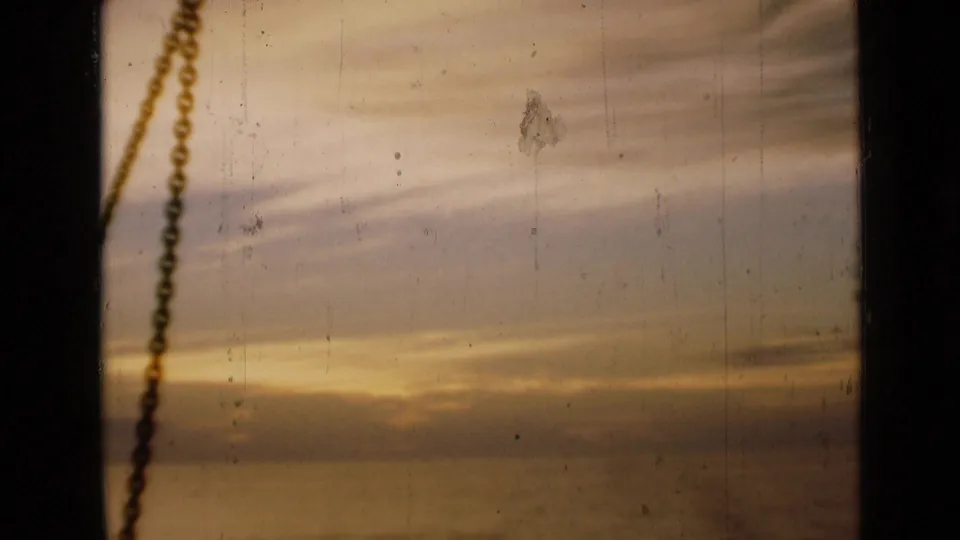 1946 London 8mm frame of a fading sunset viewed through a window, with chain silhouette, muted clouds, scratches, and dust.