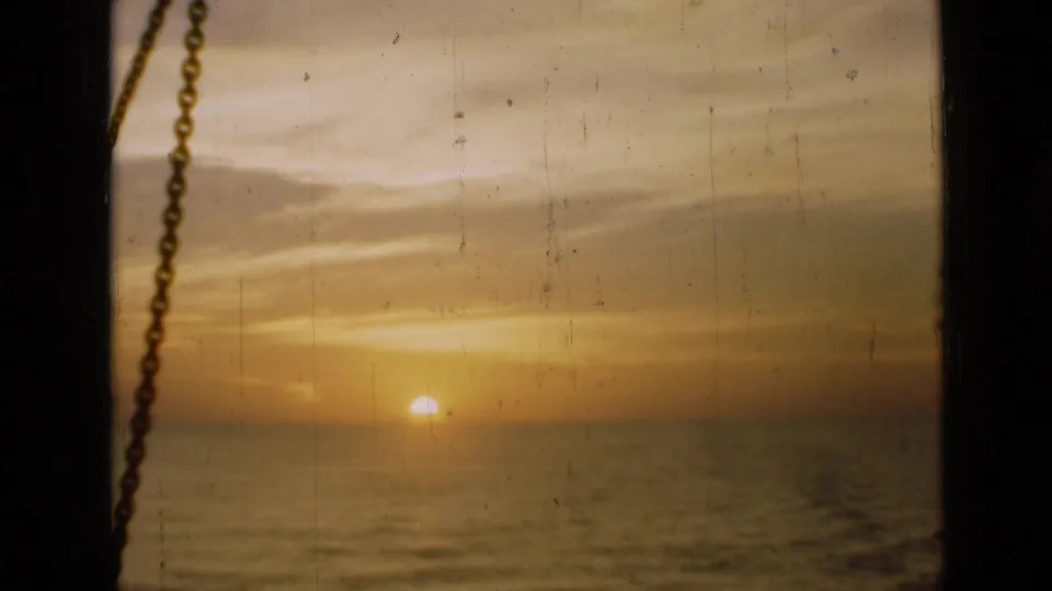 1946 8mm footage metadata places this sunset view in London, showing a low orange sun over water through a window with a chain at left.