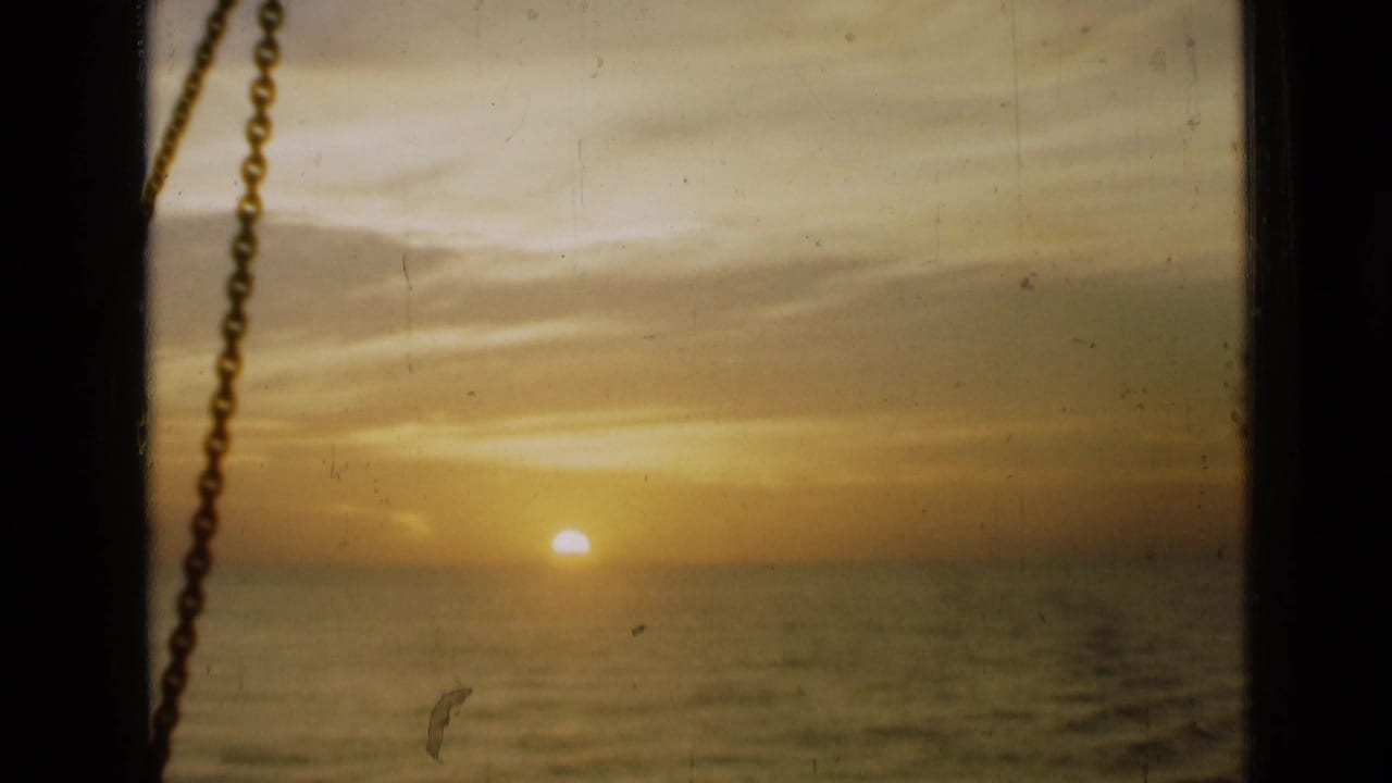 Sunset over water with clouds, viewed through a window and chain along the left side.